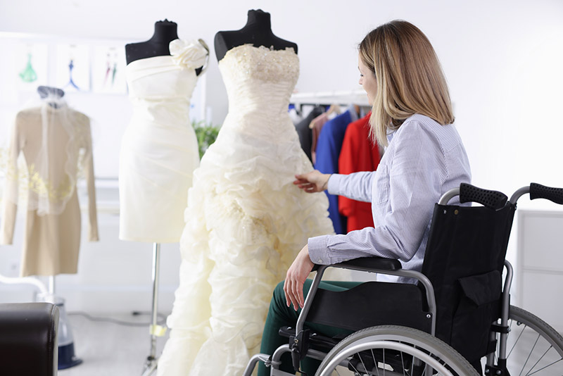 female actor seated in a wheelchair examines a wedding gown on a manniquin
