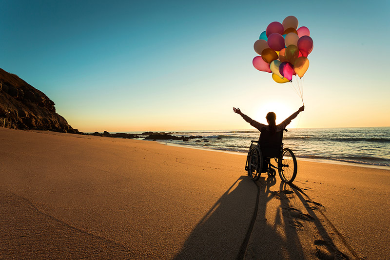 person in a hospital wheelchair on a beach holding balloons